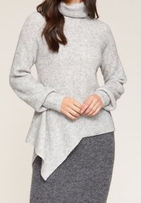 Grauer wollener Rollkragenpullover mit langen Ärmeln, der über ein strukturiertes Strickmuster und ein asymmetrisches Saumdesign verfügt, kombiniert mit einem dunkelgrauen Rock.