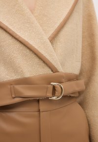 Beige tekstureret stoffrakke med et glat brunt læderbælte med en guldfarvet spænde, der fremhæver kontrasterende materialer og et sofistikeret design.