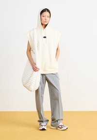 Bēša bezpiedurkņu kapuce ar melnu emblēmu, pelēkajiem platajiem biksēm un baltu teksturētu pleca soma. Sporta apavi ar bordo un baltajām akcentiem.