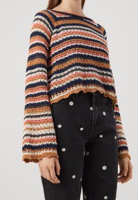 Femme portant un pull en crochet à rayures horizontales bleu marine, orange, blanc et rose, associé à un jean noir brodé de marguerites blanches.