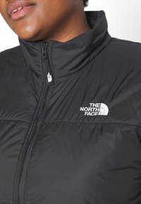 Svart zip-up jacka med hög krage, med kviltad textur och The North Face-logotyp i vitt på bröstet.