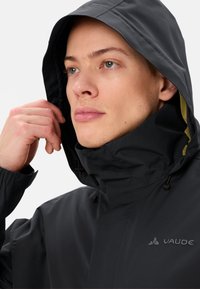 Veste noire imperméable avec capuche, fabriquée en matériau texturé. Elle dispose d'un col ajustable et d'un logo brodé discret sur la poitrine.
