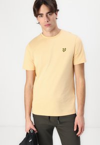 Short-sleeve, crew-neck t-shirt in light yellow cotton, featuring a small black embroidered logo on the chest. Smooth fabric texture.