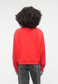 Κόκκινο φούτερ με χαλαρή εφαρμογή, ριμπ μανσέτες και στρίφωμα. Συνδυασμένο με ψηλόμεσα, ξεθωριασμένα γκρίζα τζιν. Λείο υλικό, μίνιμαλ σχεδίαση.