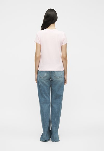 Γυναίκα με μακριά σκουρόχρωμα μαλλιά, που φοράει ανοιχτό ροζ κοντομάνικη μπλούζα και άνετο μπλε τζιν, στέκεται με την πλάτη προς τη κάμερα σε λευκό φόντο.
