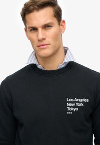 Sudadera negra con cuello de canalé, que presenta texto blanco: "Los Ángeles Nueva York Tokio." Superpuesta con una camisa de rayas azules con cuello por debajo.