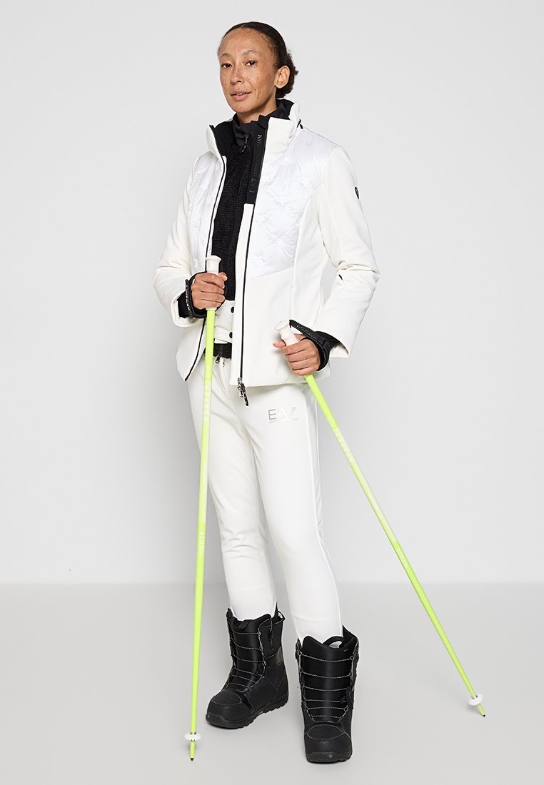 Giacca bianca trapuntata, strato interno nero, pantaloni bianchi con logo, stivali neri e bastoncini neon verdi. Tessuto liscio e vestibilità sartoriale.