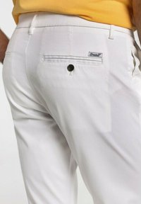 Pantalon blanc avec une poche arrière dotée d'un bouton noir et d'une étiquette. Texture lisse, coupe slim et détails de coutures subtils.