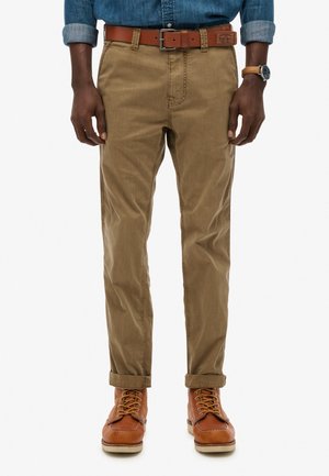 Man in opgerolde kaki broek, brown leren riem, blauwe spijkerblouse, brown leren laarzen en een horloge met een tan leren band.