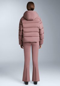 Puhasta jakna u nježno rozoj boji s kapuljačom, s prošivenim dijelovima. Nošena s pripadajućim širokim hlačama koje imaju zatvarače na gležnjevima.
