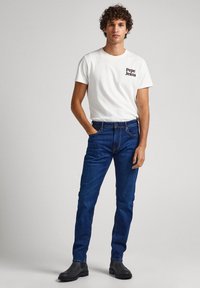 Camiseta blanca de manga corta con el logo negro de "Pepe Jeans", combinada con jeans ajustados de mezclilla azul oscuro y botas negras hasta el tobillo.