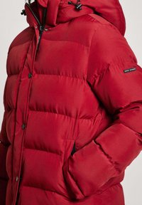 Röd puffjacka med quiltad design, som har svarta knappknappar, hög krage och en huva med snörning. Sidfickor synliga.