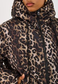 Løveprikket jakke i beige og sort, med høj krave, lynlås lukning, snore detaljer og en glat, blank tekstur.