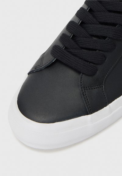 Must musta sneaker, millel on tekstuurne pind, mustad paelad, valgest kummist tald ja ümar nina. Kujundus sisaldab minimaalset detailide arvu.
