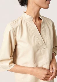 Beige, kortærmet top lavet af blødt stof med mandarin krave, der har en v-formet udskæring og subtile syninger.