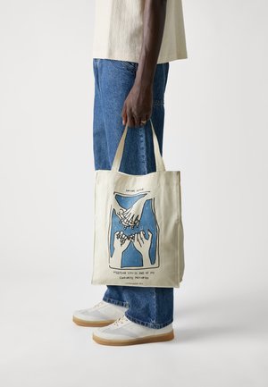 Personne portant un jean et des baskets blanches tient un sac fourre-tout beige avec une illustration bleue et noire de mains entrelaçant les petits doigts et du texte.