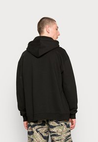 Felpa nera oversize con cappuccio ampio, texture morbida e spalle scese. Abbinata a pantaloni cargo a fantasia nei toni terrosi.