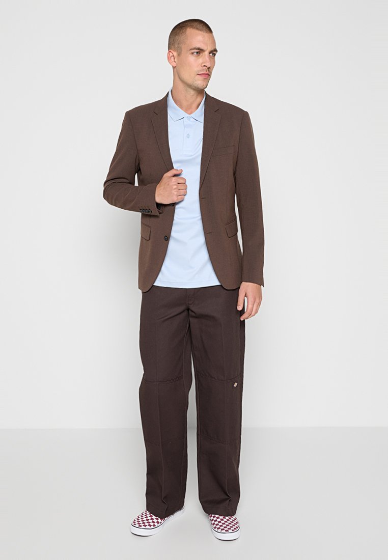 Hnědý blazer přes světle modrou polokošili, doplněný tmavě hnědými kalhotami. Outfit doplňují pruhované červenobílé tenisky. Jednoduchý, dobře padnoucí design.