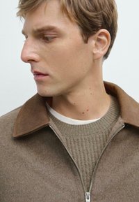 Brown zip-up jacket with a suede collar, paired with a ribbed beige sweater underneath and a white shirt. Soft texture and neutral tones.