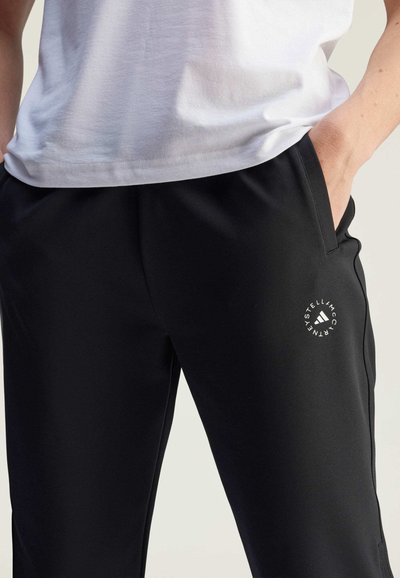 Schwarze Sporthosen mit glatter Textur, versehen mit einem kleinen weißen Logo am linken Oberschenkel und Seitentaschen. Getragen mit einem weißen Hemd.