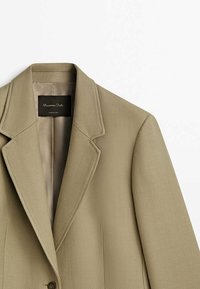 Blazer beige ajusté avec col à revers cranté, fermeture à un seul bouton, et étiquette intérieure noire avec texte blanc.