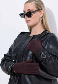 Guanti lavorati a maglia bordeaux con una finitura testurizzata, indossati su una giacca di pelle nera con grandi tasche e un accento di fibbia in argento.