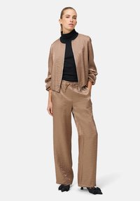 Hellbraune Satin-Bomberjacke mit überschnittenen Schultern, kombiniert mit passenden weiten Hosen und einem schwarzen Rollkragenpullover. Details des strukturierten Stoffes sind sichtbar.