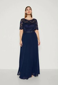 Femeie îmbrăcată într-o rochie lungă, de culoare bleumarin, cu mâneci scurte din dantelă și un brâu cu mărgele, stând în fața unui fundal deschis, simplu.