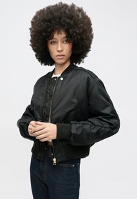 Tânăr cu păr creț purtând o geacă bomber neagră și blugi închiși la culoare, stând în picioare pe un fundal deschis, cu mâinile împreunate.