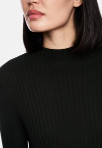 Schwarzer gerippter Pullover mit Stehkragen, der feine vertikale Linien und eine glatte Textur aufweist. Nahaufnahme des Kragens und des oberen Brustbereichs.