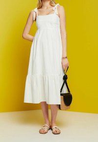 Femme portant une robe midi blanche sans manches avec des bretelles nouées, des sandales blanches, et tenant un sac à main tressé avec des accents en cuir noir.