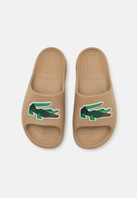 Bruine rubberen slidesandalen met een gestructureerde voetbed, open teen en een groen geborduurd krokodillenlogo op de bovenkant.