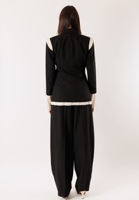 Donna con lunghi capelli scuri che indossa pantaloni neri a gamba larga e un blazer nero con dettagli bianchi, in piedi di fronte a uno sfondo bianco semplice.