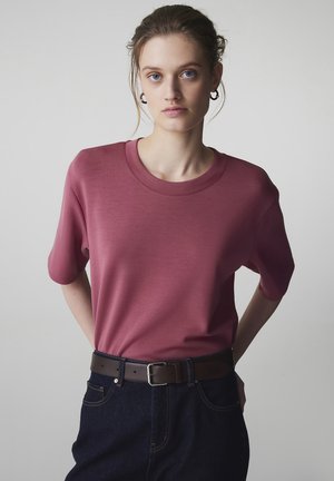 Jonge vrouw met lichte huid en blauwe ogen draagt een donkerroze T-shirt met korte mouwen, donkere spijkerbroek, bruine riem en creolen tegen een grijze achtergrond.