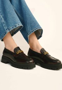 Burgundowe skórzane mokasyny z złotym łańcuszkiem, masywną czarną podeszwą i gładką fakturą. W połączeniu z dżinsami o szerokich nogawkach.