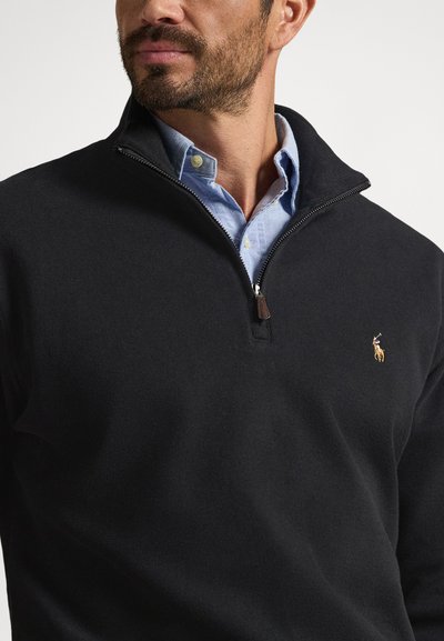 Suéter preto com fecho zip, confeccionado em tecido de malha, apresenta um pequeno logotipo bordado e é usado sobre uma camisa azul claro com colarinho.