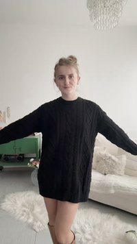 Mladá žena stojí v interiéri s rozpaženými rukami, oblečená v čiernom svetrovom šate s vrkočovým vzorom, v pozadí je biela pohovka, zelená skrinka a lustre.