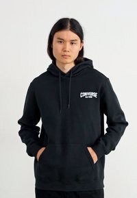 Sudadera negra con capucha, bolsillo frontal, cordones y logo "Converse All Star" en blanco en el pecho. Textura suave, diseño casual.