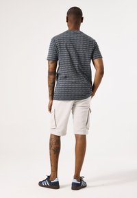 Camisa de manga corta a rayas grises, pantalones cortos cargo blancos y zapatillas azules con acentos negros. Presenta tatuajes visibles en la parte inferior de la pierna.