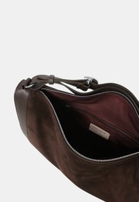 Brązowa torebka z zamszu i skóry z srebrnym zamkiem błyskawicznym, otwarta, aby pokazać ciemną tkaninę wewnętrzną oraz wewnętrzną kieszeń na zamek z metką marki.