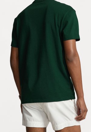 Mann trägt dunkelgrünes kurzärmeliges Poloshirt und weiße Shorts, von hinten vor einem schlichten hellen Hintergrund gezeigt.