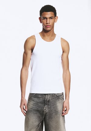 Jeune homme portant un débardeur blanc uni sans manches et un jean gris délavé, debout devant un fond blanc.