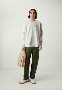 Young man standing with neutral expression wearing white sweatshirt, dark green pants, white sneakers, and holding beige backpack by side.