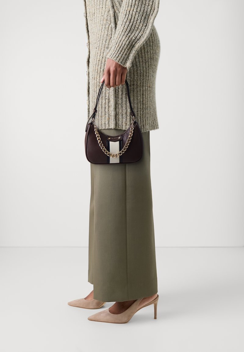 Personne en pull en maille beige et pantalon olive tenant un petit sac à main marron foncé avec détail chaîne, portant des talons hauts beiges.