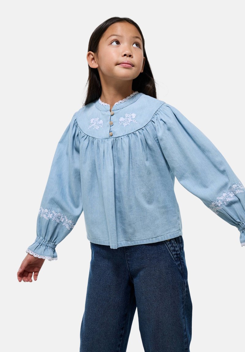 Blouse en denim bleu clair avec des accents floraux brodés, corsage froncé et longues manches bouffantes, dotée d'une fermeture à boutons et d'un bord en dentelle.