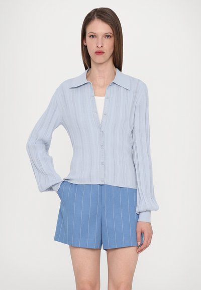 Lyseblå strikket cardigan med ribbet tekstur, knappelukking og spiss krage, sammen med blå stripete shorts.