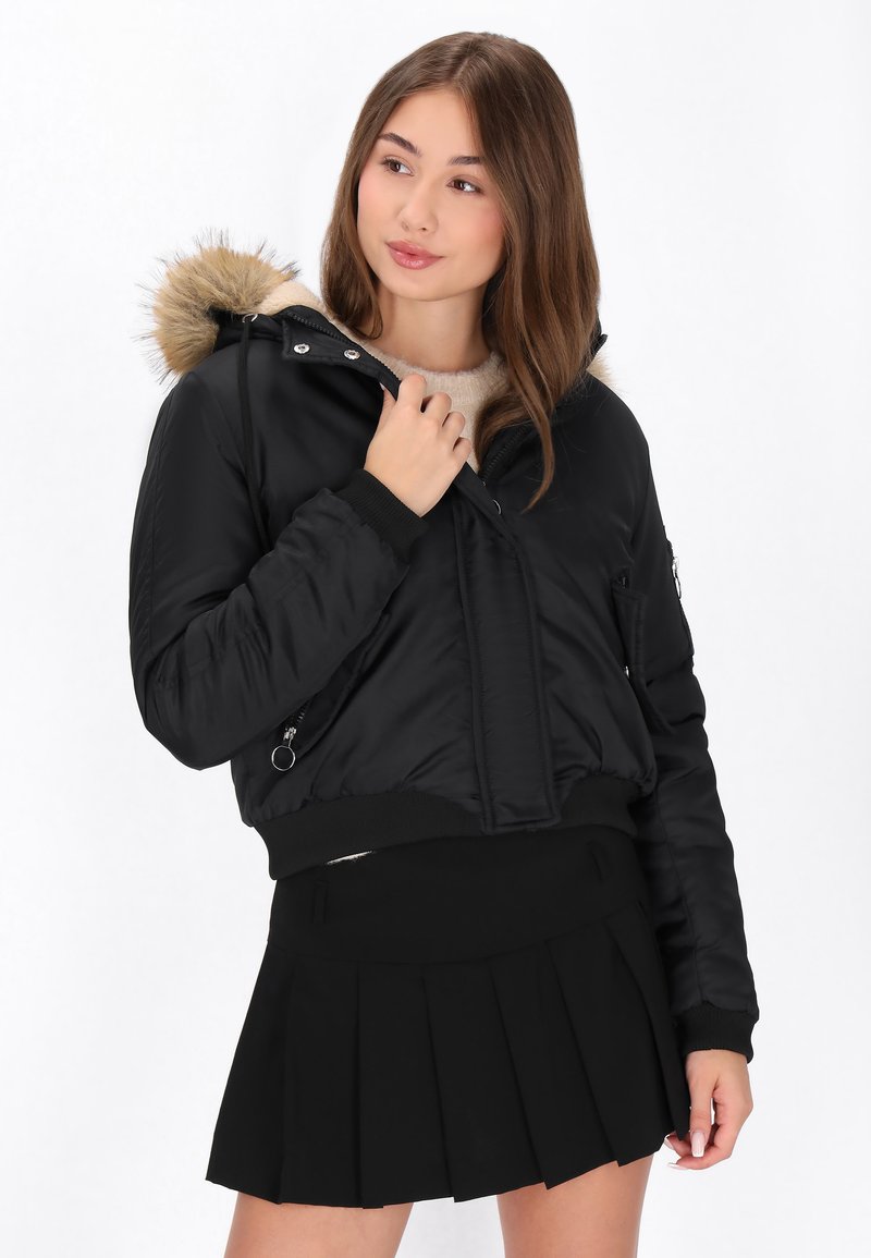 Sort bomberjakke med en faux fur hætte, med lynlås lukning, sidelommer, ribbede manchetter og et cropped design, båret over en plisseret sort nederdel.
