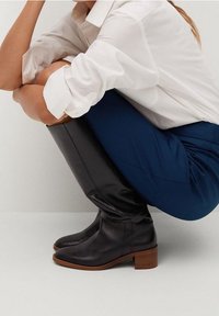 Bottes en cuir noir hautes avec un bout arrondi et un talon moyen empilé, portées avec une chemise blanche et un pantalon bleu, mettant en valeur une texture lisse.
