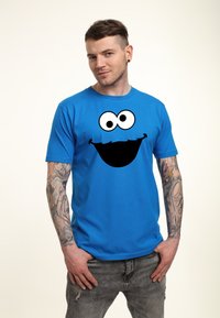 Modrá bavlněná tričko s velkým černým grafikem usměvavé tváře s velkýma očima a širokým úsměvem. Model má tetování a nosí šedé džíny.
