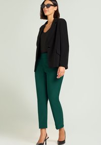Zwarte blazer met een enkele knoop, groene aansluitende broek en zwarte slingback hakken. De outfit bevat een donker topje en moderne zonnebril.